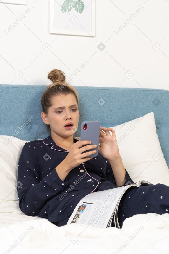 Primo piano di una giovane donna scioccata in pigiama sdraiata a letto mentre naviga in rete
