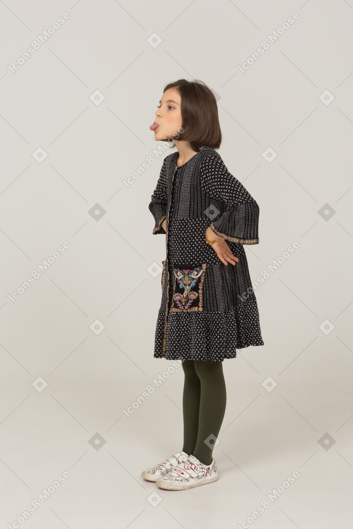 Трехчетвертный вид забавной маленькой девочки в платье, показывающей язык и кладущей руки на бедра