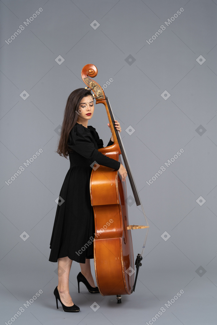 Вид сбоку на серьезную молодую музыкантку в черном платье, играющую на контрабасе