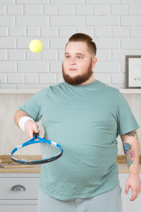 A fat man holding a tennis racket and playing with a tennis ball