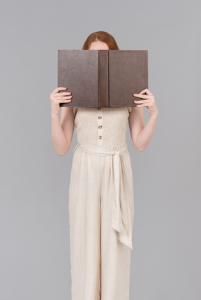 Teenage girl dressed in beige overalls closing face with the book