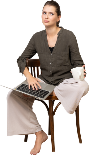 Front view of a confused young woman wearing home clothes sitting on a chair with a laptop & coffee cup