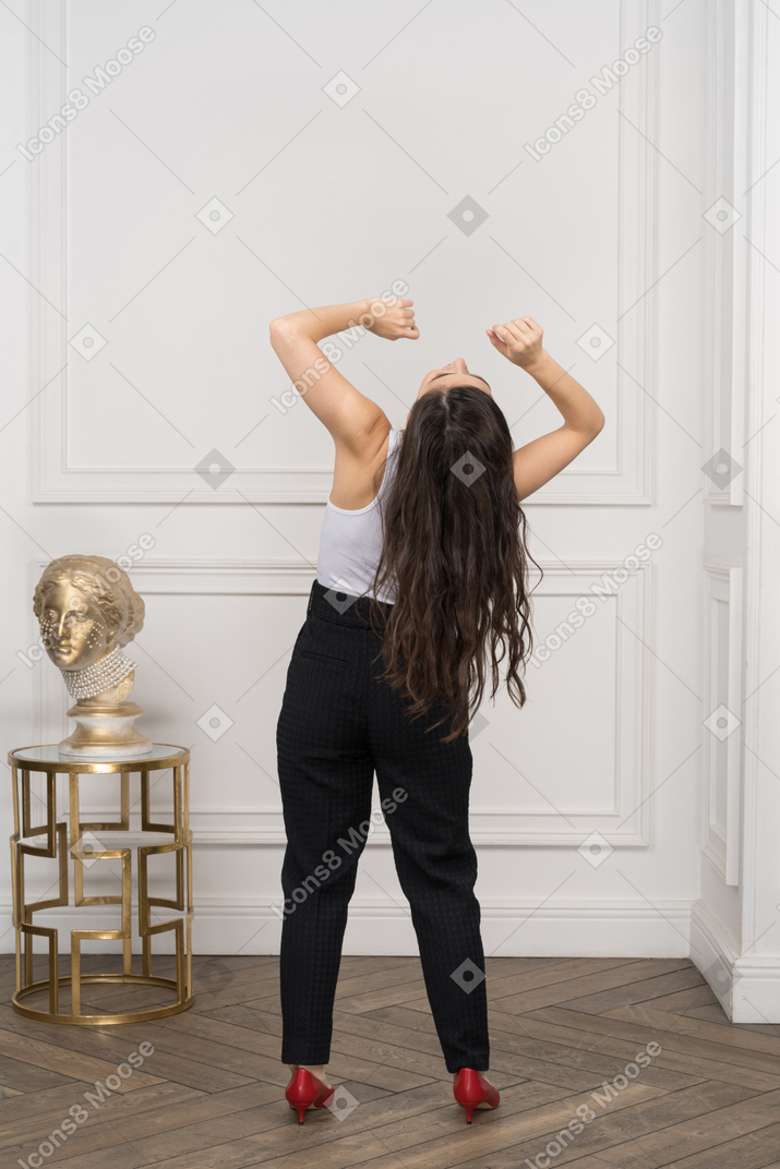 Back view of a young female raising hands and throwing head back while standing by a golden greek sculpture