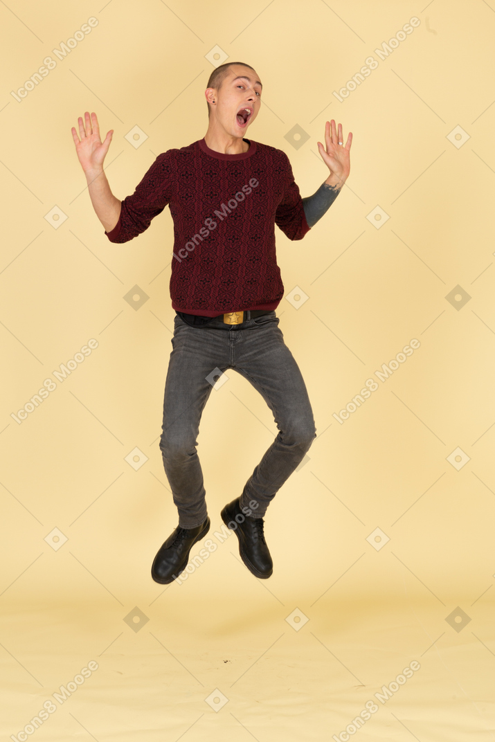 Front view of a jumping young man in red pullover raising his hands