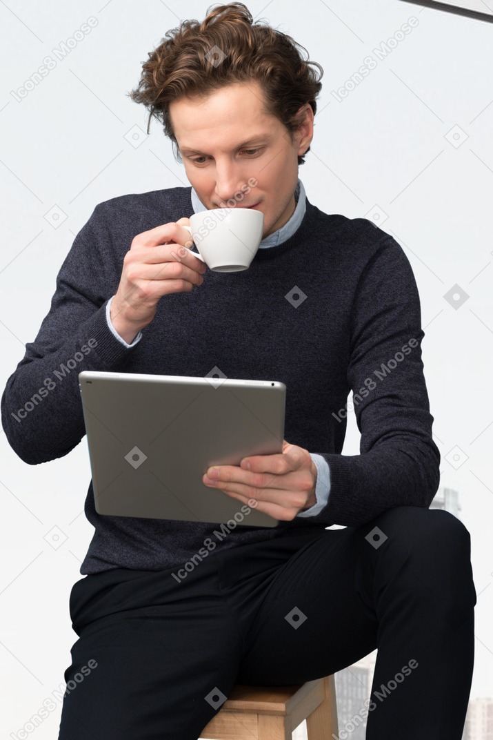 A man sitting on a stool drinking a cup of coffee while using a tablet