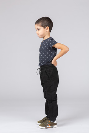 Vista lateral de um lindo menino em roupas casuais, posando com a mão nas costas e fazendo caretas