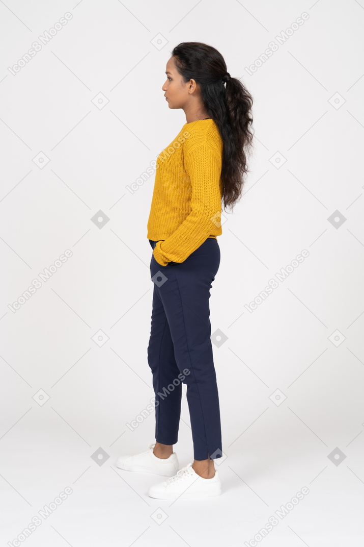 Vista lateral de una niña en ropa casual posando con las manos en los bolsillos