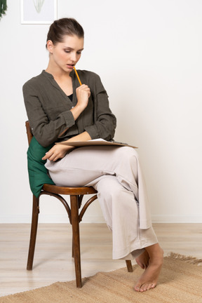 Vue de face d'une jeune femme réfléchie portant des vêtements de maison assise sur une chaise et prenant des notes
