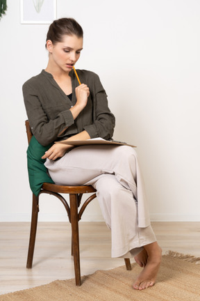 Front view of a thoughtful young woman wearing home clothes sitting on a chair and making notes