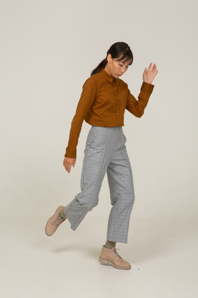 Vista di tre quarti di una giovane donna asiatica in calzoni e camicetta che alza la mano e alza la gamba