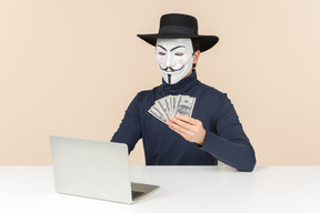 Hacker vestindo máscara de vingança sentado à mesa e contando dinheiro