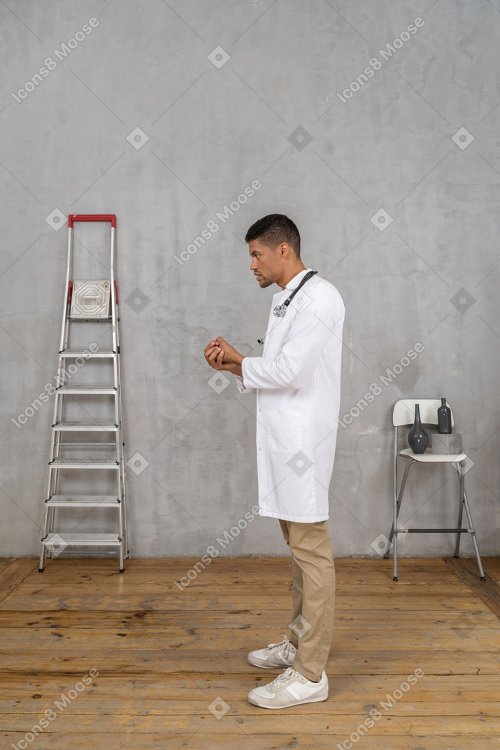 Side view of a young doctor standing in a room with ladder and chair and holding hands together