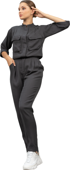 Front view of a walking young woman in a jumpsuit holding hand in pocket & touching head  download