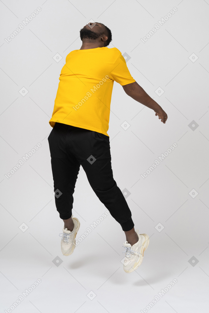Vista de três quartos de um homem jovem de pele escura pulando em uma camiseta amarela estendendo as mãos