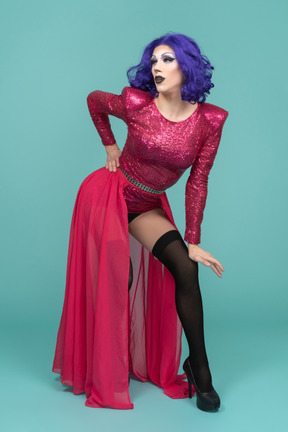 Retrato de uma drag queen de vestido rosa posando com as costas arqueadas e a mão apoiada no joelho