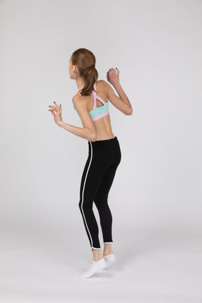 Vista traseira de três quartos de uma adolescente em roupas esportivas levantando as mãos enquanto pula