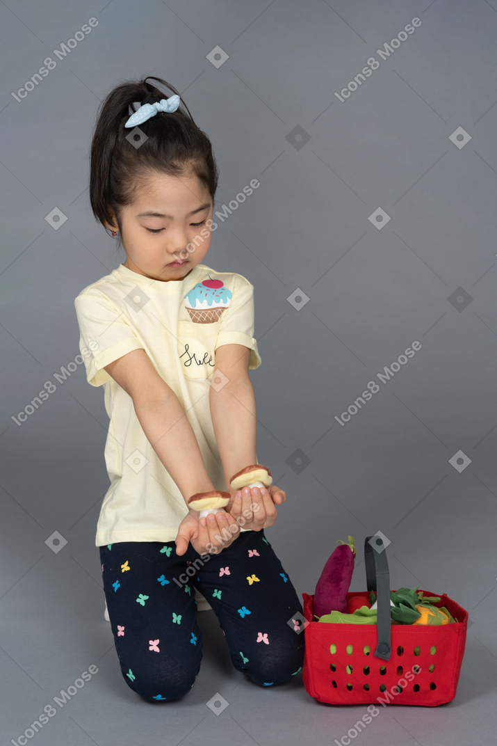 Little girl holding two mushrooms while sitting beside a shopping basket