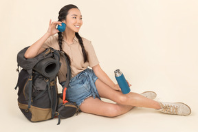 Lächelnde junge asiatische frau, die nahe rucksack sitzt und thermosflasche hält