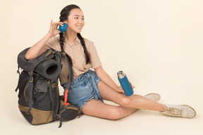 Sonriente joven asiática sentada cerca de mochila y sosteniendo termo