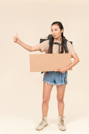 Young female hitchhiker holding paper card and showing thumb up