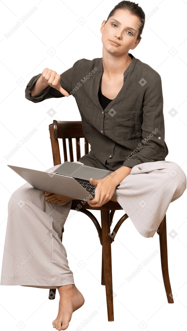 Front view of a displeased young woman sitting on a chair with a laptop & showing thumb down