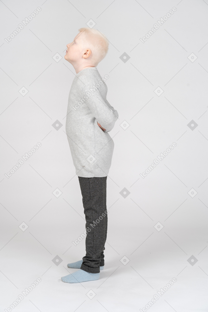 Um garotinho loiro em roupas casuais em pé e escondendo as mãos atrás