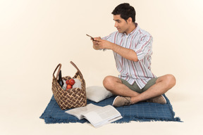 Молодой кавказский парень сидит на одеяле и делает фотографию корзины для пикника