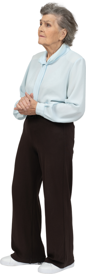 Vista frontale di una donna anziana seria in camicetta e pantaloni che tengono le mani insieme