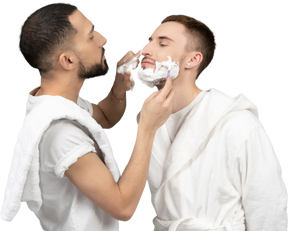 Young caucasian man carefully shaving his partner and putting shaving foam on his nose
