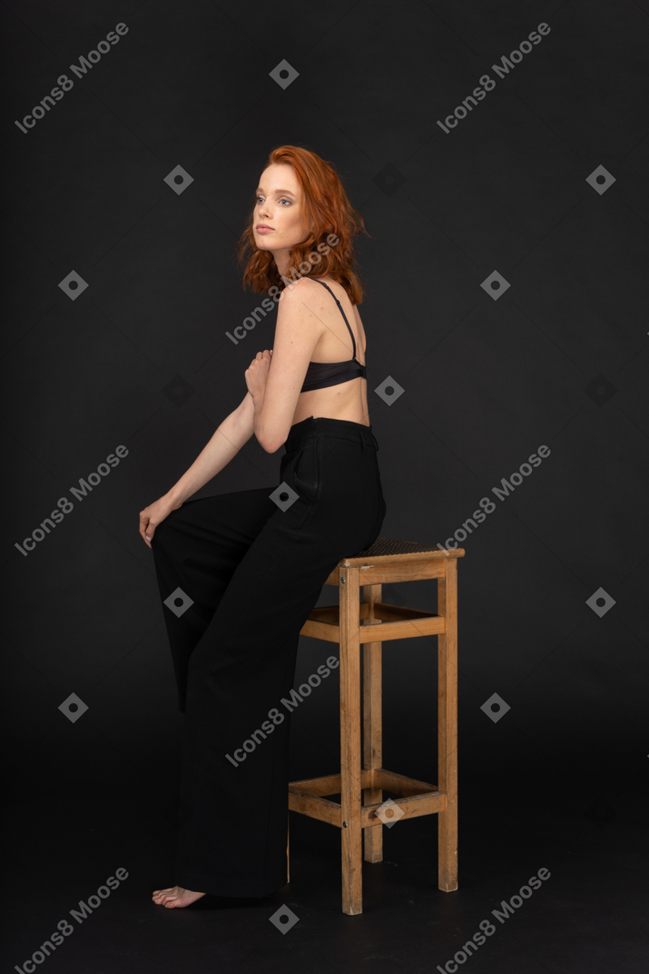 A side view of the beautiful woman dressed in black pants and bra, sitting on the wooden chair and touching her hand
