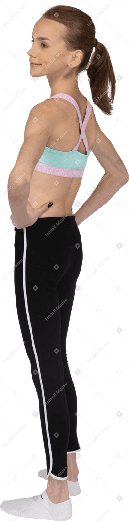 Vista posteriore di tre quarti di una ragazza adolescente in abbigliamento sportivo che mette le mani sui fianchi sorridendo leggermente