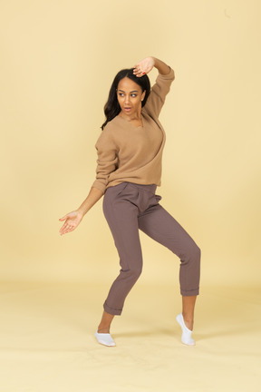 Вид в три четверти темнокожей танцующей молодой женщины, поднимающей руку, глядя в сторону