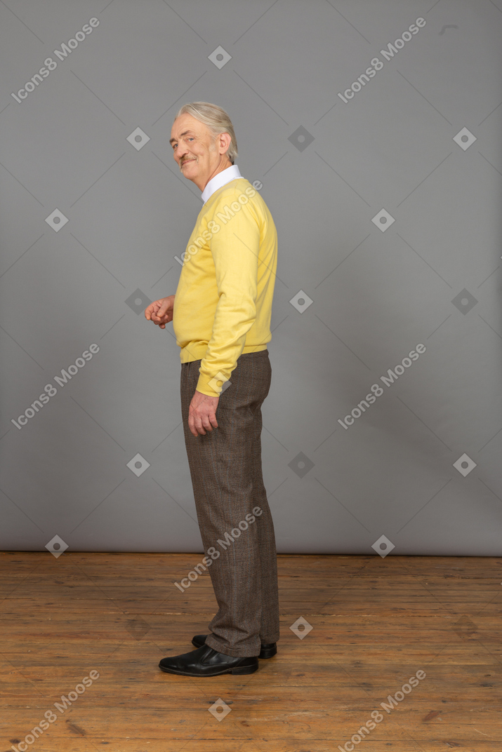 Side view of a smiling old man in yellow pullover raising hand and looking at camera