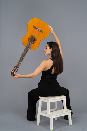 Back view of a young lady in black suit holding the guitar upside down and sitting on stool