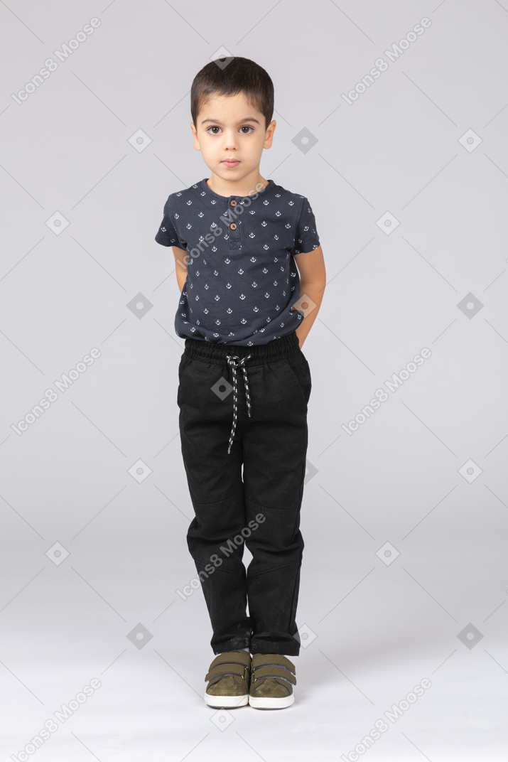 Front view of a cute boy standing with hands behind back and looking at camera
