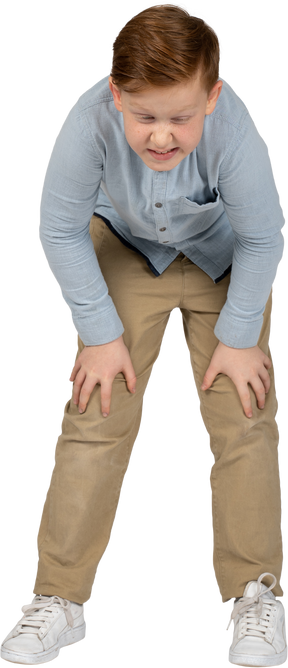 Vista frontal de un niño agachándose y tocando las rodillas lastimadas