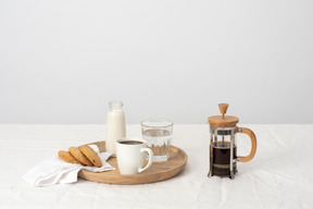 Café en prensa francesa y taza grande de café, vaso de agua, leche y galletas en la bandeja.