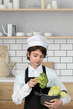 A young boy in a chef's outfit holding a pot of vegetables