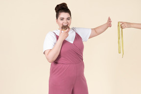 Young plus size woman in a fuchsia jumpsuit posing with food against a pastel yellow background