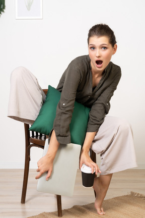 Vista frontale di una giovane donna scioccata seduta su una sedia e tenendo il suo laptop e toccando la tazza di caffè