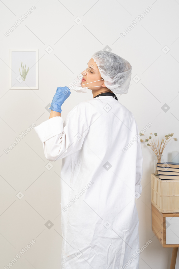 Three-quarter back view of a displeased young female doctor trying to take mask off