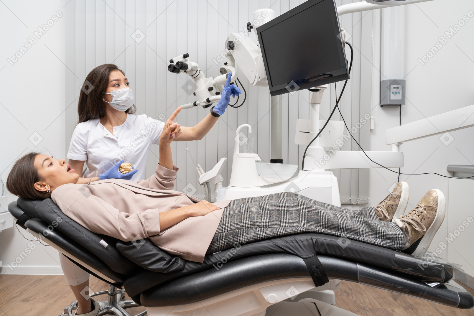 Женщина-стоматолог в полный рост консультируется со своей пациенткой, одновременно указывая на экран