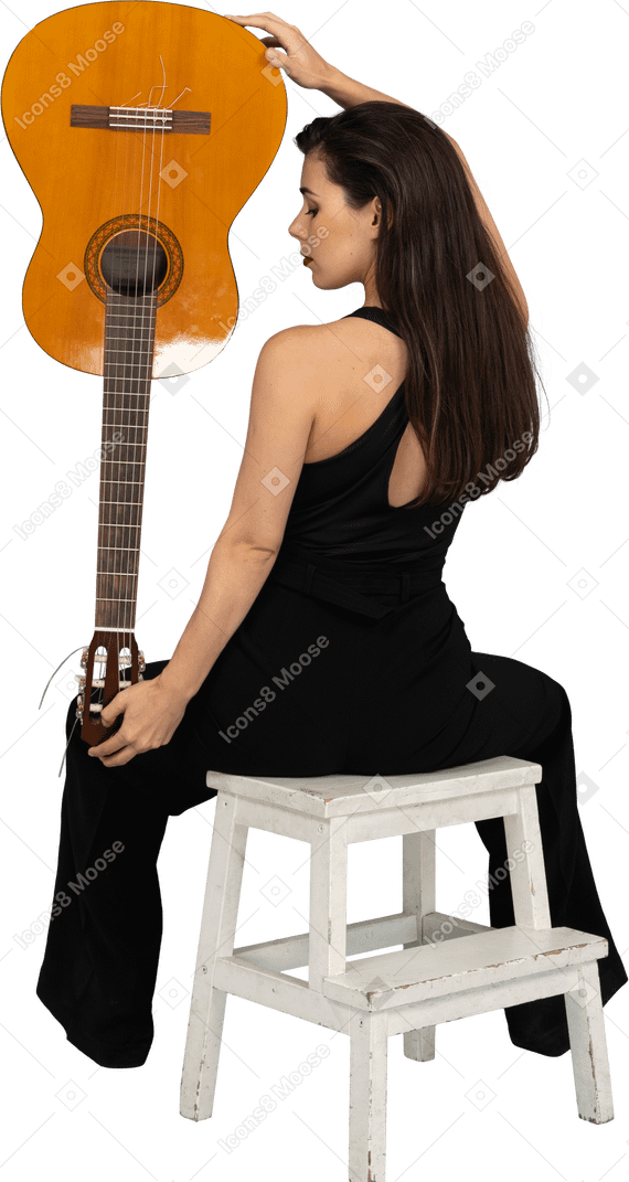 Back view of a young lady in black suit holding the guitar upside down and sitting on stool