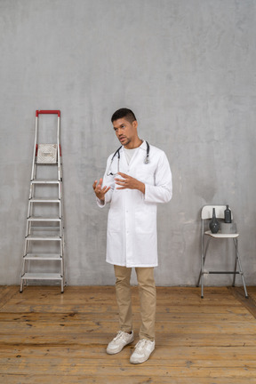 Vista de tres cuartos de un joven médico gesticulante de pie en una habitación con escalera y silla