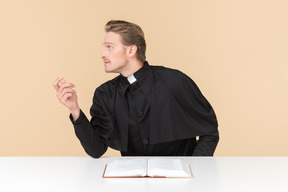 Catholic priest sitting at the table with open bible book and like saying something