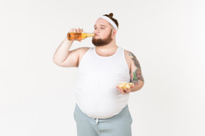 A fat man in sportswear drinking beer and holding a handful of chips