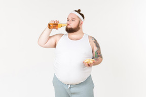 Um homem gordo no sportswear bebendo cerveja e segurando um punhado de batatas fritas