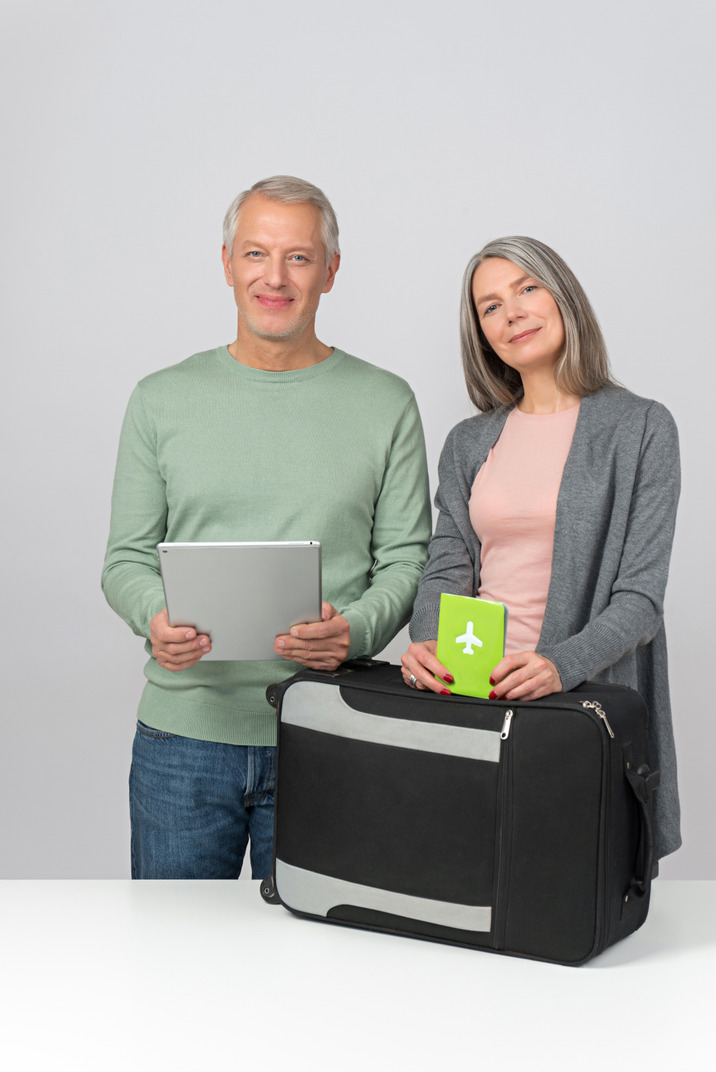 Middle aged couple holding tablet and passport standing next to suitcase