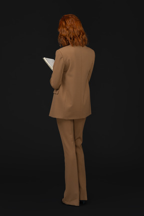 Standing back to camera young woman in formal suit writing something down in notebook