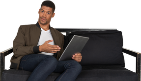 Front view of a shocked young man sitting on a sofa while watching the tablet